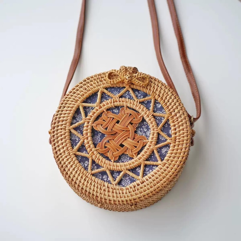 K&ouml;nnte beinhalten: Eine runde, gewebte Korbtasche mit einem braunen Lederriemen. Die Tasche hat ein blau gemustertes Stofffutter und ein dekoratives Webdesign in der Mitte. Die Tasche hat eine nat&uuml;rliche, beige Farbe.