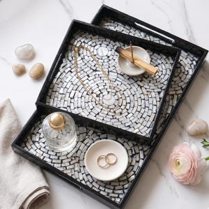 May include: Three black decorative trays with a mosaic tile design. The trays hold a gold necklace, a small dish with two gold rings, a perfume bottle, and a small dish with a stick of incense. Decorative stones and a pink flower are also present.