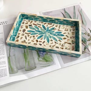 Pode incluir: Uma bandeja decorativa retangular com um design de mosaico. A bandeja apresenta um padr&atilde;o floral turquesa e branco no centro, com detalhes turquesa nas bordas. A bandeja est&aacute; sobre uma revista.