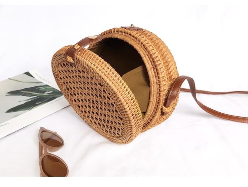K&ouml;nnte beinhalten: Eine runde, gewebte Korbtasche mit einem braunen Lederriemen und Griff. Die Tasche ist ge&ouml;ffnet und zeigt ein Stofffutter. Braune Sonnenbrille und eine Zeitschrift liegen neben der Tasche.