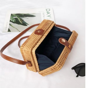 K&ouml;nnte beinhalten: Rechteckige Umh&auml;ngetasche aus geflochtenem Rattan mit braunem Lederriemen und Akzenten. Die Tasche hat einen Klappverschluss und ein dunkelblaues Innenfutter. Eine Sonnenbrille ist sichtbar.