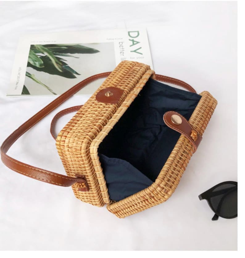 K&ouml;nnte beinhalten: Rechteckige Umh&auml;ngetasche aus geflochtenem Rattan mit braunem Lederriemen und Akzenten. Die Tasche hat einen Klappverschluss und ein dunkelblaues Innenfutter. Eine Sonnenbrille ist sichtbar.
