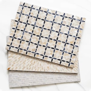 May include: Three rectangular placemats featuring distinct surface patterns. The top mat displays a dark blue four-petal floral grid on a cream shell background. The middle mat has a radial leaf-like pattern, and the bottom mat shows a light grey brick-like mosaic design.