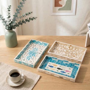 May include: Three rectangular decorative trays with mosaic designs. One tray features a blue floral pattern, another has a beige leaf pattern, and the third showcases a fish motif in blue and various colors. A cup of coffee sits on a beige placemat.