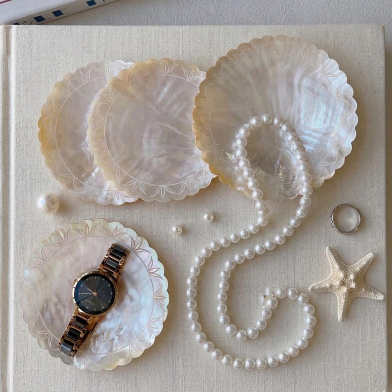 K&ouml;nnte beinhalten: Eine Anordnung dekorativer Gegenst&auml;nde, darunter vier schillernde Muschelschalen-Untersetzer mit gewellten R&auml;ndern, eine Perlenkette, eine Armbanduhr, ein Silberring und ein Seestern. Auf einer neutralen Oberfl&auml;che angeordnet.