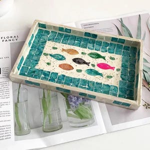 Pode incluir: Bandeja decorativa retangular com borda de mosaico turquesa. O centro da bandeja apresenta um desenho de peixes coloridos. A bandeja est&aacute; sobre uma revista com o texto "FLORAL FANCY".