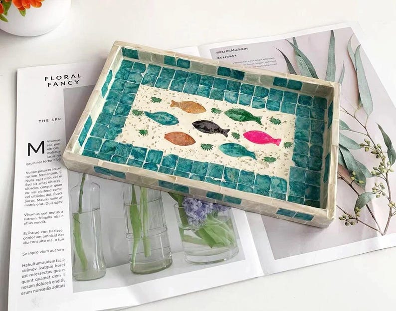 Pode incluir: Bandeja decorativa retangular com borda de mosaico turquesa. O centro da bandeja apresenta um desenho de peixes coloridos. A bandeja est&aacute; sobre uma revista com o texto "FLORAL FANCY".