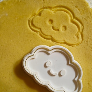 Può includere: Un tagliabiscotti bianco a forma di nuvola con una faccia sorridente, accanto a un'impronta dello stesso disegno in pasta gialla. Il tagliabiscotti ha un bordo arrotondato ed è posto sulla pasta, pronto per l'uso. La pasta è di colore giallo chiaro.