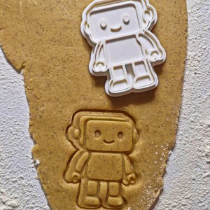 May include: A white plastic robot-shaped cookie cutter sits on a piece of dough, with an impression of the robot cut into the dough below. The dough is light brown, and white flour is sprinkled around the edges.
