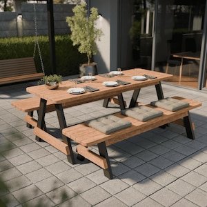 May include: A wooden picnic table with attached benches. The table has a light brown finish with black legs. The benches have gray cushions. Table settings include plates, silverware, and glasses. A small potted plant sits on the table.