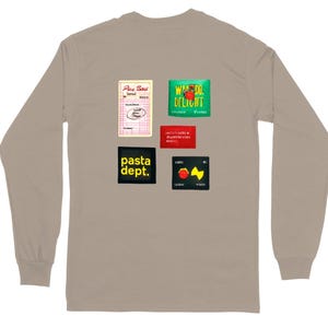 May include: A long-sleeve taupe shirt with several colorful graphic patches. The patches include text and illustrations, such as "Pasta Dept." and a tomato. The shirt has a crew neck and long sleeves.