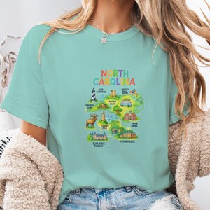 Peut inclure: T-shirt bleu clair avec une carte colorée de la Caroline du Nord, ornée de monuments et du texte "NORTH CAROLINA". Le design comprend des illustrations du phare de Cape Hatteras et du Tryon Palace. Col rond, manches courtes.