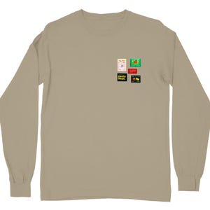 May include: A long-sleeve, tan-colored shirt with a crew neck. The shirt features a small collection of colorful graphic designs on the upper left chest. The graphics include text and illustrations.