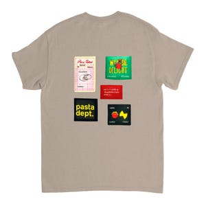 May include: A tan t-shirt with several colorful patches. The patches include text and graphics such as "Pasta Dept.", "Wardo Delight", and a red tomato with a yellow lightning bolt.