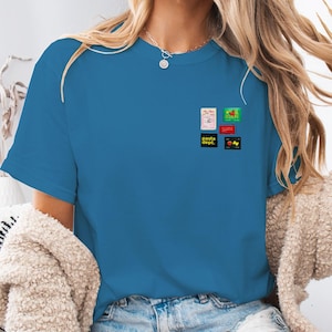 Peut inclure: Un t-shirt bleu turquoise avec plusieurs petits motifs graphiques colorés sur la poitrine. Les motifs comprennent du texte et des illustrations liés aux pâtes. Le texte "pasta dept." est également visible.