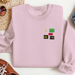 May include: A light pink sweatshirt with a crew neck. The sweatshirt features several small, colorful graphic designs, including the text "pasta dept." and other food-related imagery. The design is located on the upper left chest area.