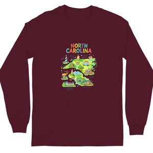 Peut inclure: Un t-shirt à manches longues bordeaux avec une carte colorée de la Caroline du Nord. La carte comprend des illustrations de monuments et d'icônes, et le texte "NORTH CAROLINA" en arc-en-ciel.