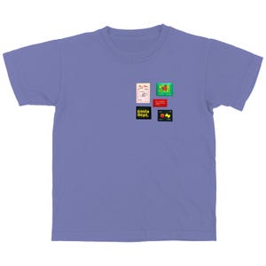 May include: A light purple t-shirt with a crew neck and short sleeves. The shirt features several small, colorful rectangular graphics on the upper left chest area. The graphics include text and illustrations related to pasta and Italian cuisine.