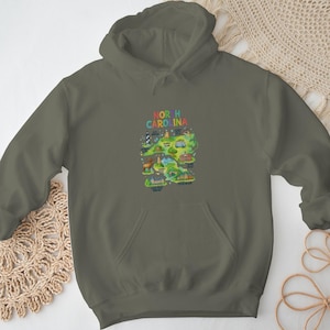 Peut inclure: Sweat à capuche vert olive avec une carte colorée de la Caroline du Nord. La carte comprend des illustrations de monuments et le texte "NORTH CAROLINA" en couleurs arc-en-ciel. Le sweat à capuche a une poche avant et une capuche à cordon.