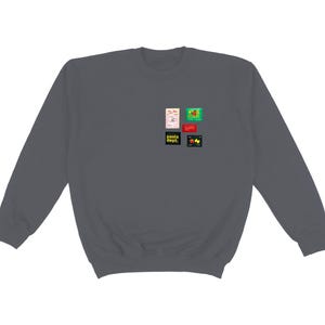 May include: A dark gray sweatshirt with a crew neck. The sweatshirt has several small, colorful rectangular patches on the upper left chest. The patches feature various designs and text, including the words "pasta dept."