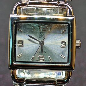 May include: A silver-toned rectangular quartz watch with a light-colored face. The watch has a silver band and a clear display stand. The watch face displays the numbers 3 and 9, and the word "QUARTZ".