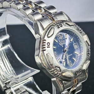 May include: Silver-toned wristwatch with a blue dial and a date display. The watch features a stainless steel band and a round bezel with numerals. The watch is displayed on a clear stand.