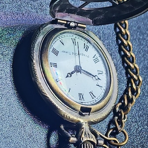 May include: Antique pocket watch with a bronze-colored case and chain. The watch face displays Roman numerals and the words "Prague Old Timer." The watch is open, revealing the time. The background is a dark, textured surface.