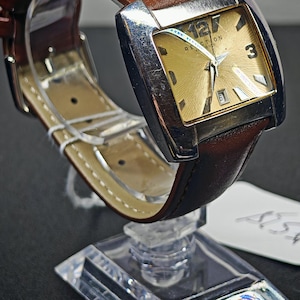 May include: A rectangular wristwatch with a gold-colored face and silver-toned bezel. The watch has a brown leather band. The watch face displays the numbers 12 and 3. The watch is displayed on a clear stand.
