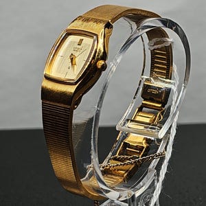 May include: A gold-toned Citizen Quartz watch with a rectangular face and a mesh band. The watch is displayed on a clear stand, showcasing its elegant design and vintage appeal. The watch face displays the Citizen Quartz logo.