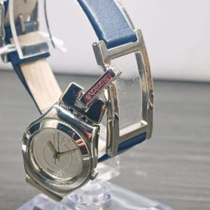 May include: A silver-tone wristwatch with a round face, featuring a white dial with silver hands and a prominent number 12. The watch has a blue leather strap and a metallic silver-tone frame. The word Swatch is printed on a small red rectangular tab.