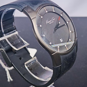 May include: A black Kenneth Cole New York watch with a dark gray face and a black rubber strap. The watch has a date display and a red second hand. The watch is displayed on a clear stand.