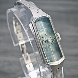 May include: A silver-toned Seiko wristwatch featuring a rectangular case and a matching metal link bracelet. The watch face is a dark, gradient green with thin silver hour markers and hands. A small crown is positioned on the right side of the case.