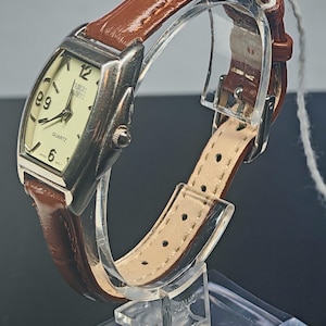 May include: A rectangular quartz watch with a silver-tone case and a brown leather strap. The watch face is cream-colored with black numerals and hands. The watch is displayed on a clear acrylic stand.