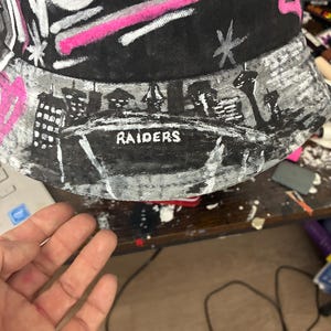 May include: A black bucket hat featuring hand-painted white and pink designs. The brim displays a detailed black and white cityscape silhouette with the word RAIDERS printed in white. The crown includes white script text, pink heart accents, and star motifs.