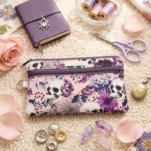 May include: A rectangular fabric pencil case featuring a pink background with a pattern of white skulls, purple flowers, and spiderwebs. The case has two zippered compartments, a metal D-ring on the side, and a label reading Sabelle Design Studio.