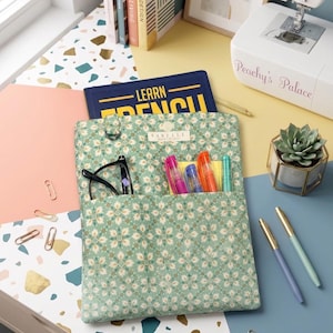 May include: A rectangular fabric pouch featuring a light green floral pattern with a quilted texture. The pouch has two front pockets containing black eyeglasses and four colorful pens. A label reading Sabelle is centered at the top edge.