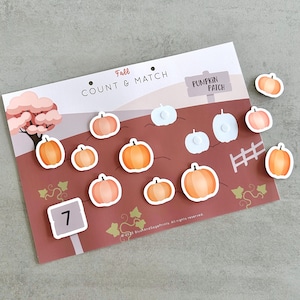 Op de afbeelding: Een educatief spel met herfstthema met de tekst "Fall Count & Match" en "Pumpkin Patch". Het spel bevat oranje pompoenvormige magneten en een kaart met een pompoenveldscène. Een bord toont het nummer 7.