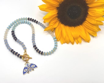 May include: A beaded necklace with a blue and gold moth pendant. The necklace features a mix of blue, white, and black beads. A bright yellow sunflower is in the background.