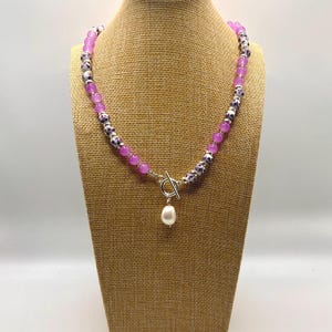 May include: A beaded necklace with a toggle clasp and a dangling pearl pendant. The necklace features alternating purple and patterned beads. The necklace is displayed on a mannequin.