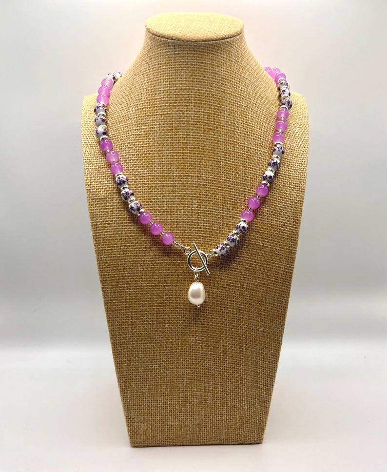 May include: A beaded necklace with a toggle clasp and a dangling pearl pendant. The necklace features alternating purple and patterned beads. The necklace is displayed on a mannequin.