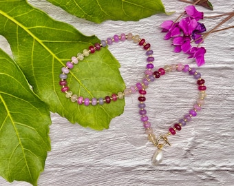 May include: A beaded necklace with a gold clasp and a dangling pearl pendant. The necklace features a gradient of pink, purple, and yellow beads. The necklace is displayed on a green leaf with pink flowers in the background.