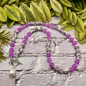 May include: A necklace featuring a strand of vibrant purple beads interspersed with white beads adorned with a floral pattern. The necklace is secured with a silver toggle clasp and a dangling pearl pendant. The jewelry is displayed against a brick background.