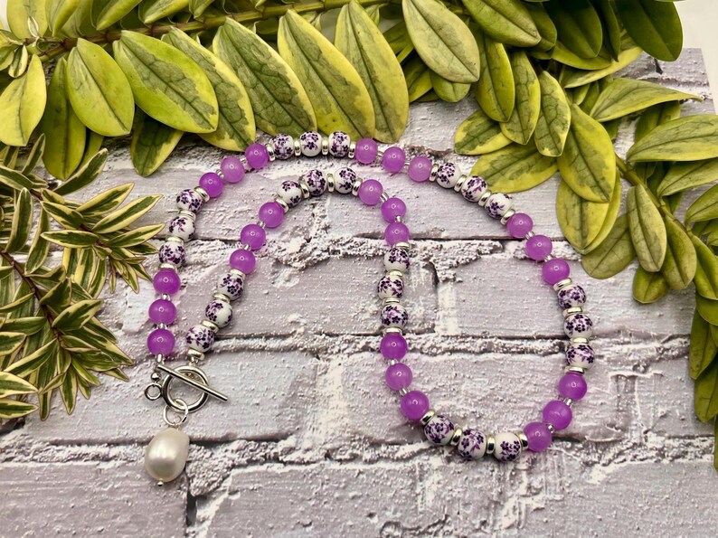 May include: A necklace featuring a strand of vibrant purple beads interspersed with white beads adorned with a floral pattern. The necklace is secured with a silver toggle clasp and a dangling pearl pendant. The jewelry is displayed against a brick background.