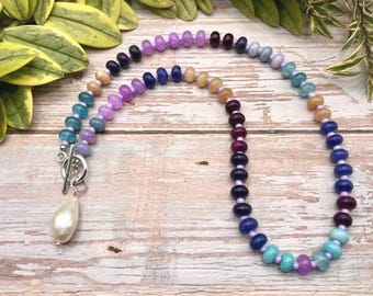 May include: A colorful beaded necklace with a pear-shaped pendant. The necklace features a variety of beads in shades of blue, purple, orange, and brown. The pendant is a large, white, teardrop-shaped pearl. The necklace is secured with a silver clasp.