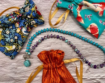 May include: A turquoise and purple beaded necklace with a silver clasp and a turquoise pendant. The necklace is displayed with three fabric pouches in blue floral, teal koi fish, and orange geometric patterns, each with a drawstring closure.