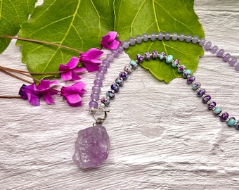 May include: A handcrafted necklace featuring a large, raw amethyst pendant. The necklace incorporates lavender-colored beads and decorative beads with purple and turquoise accents. Bright pink flowers and green leaves provide a natural backdrop.