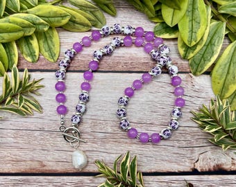 May include: A purple beaded necklace with a toggle clasp and a white, teardrop-shaped pearl pendant. The necklace features alternating sections of solid purple beads and white beads with a purple floral pattern. The necklace is displayed on a wooden surface.