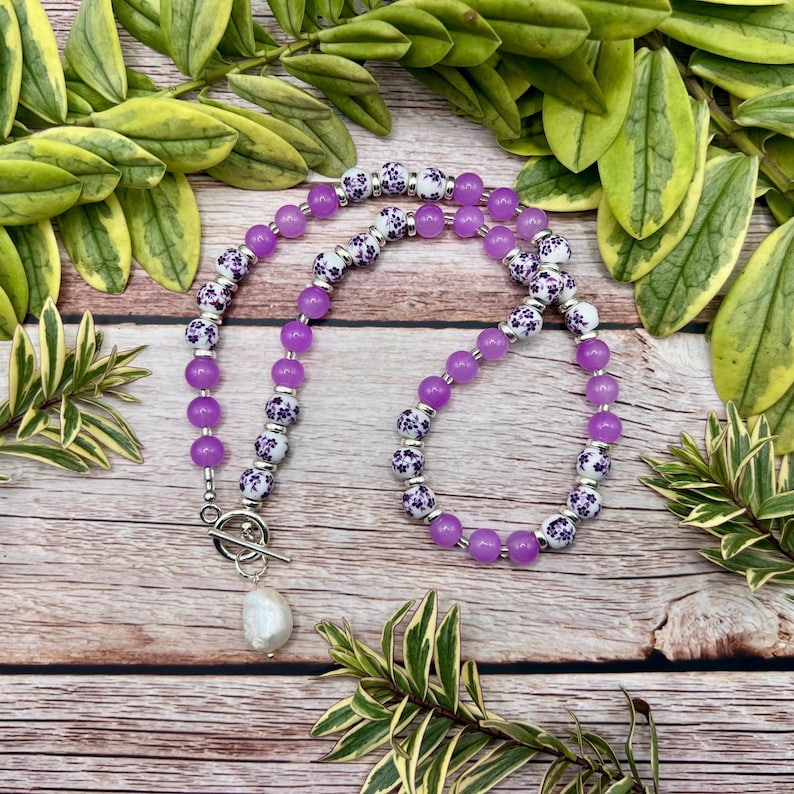 May include: A purple beaded necklace with a toggle clasp and a white, teardrop-shaped pearl pendant. The necklace features alternating sections of solid purple beads and white beads with a purple floral pattern. The necklace is displayed on a wooden surface.