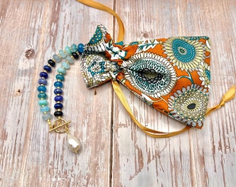May include: A beaded necklace with shades of blue and gold clasp, next to a patterned orange fabric pouch with a floral design. The necklace features a large pearl pendant. The pouch has a bow shape and yellow ribbons.