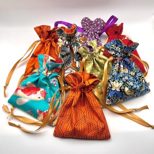 May include: A collection of colorful drawstring pouches in various patterns. The pouches feature designs such as koi fish, floral prints, polka dots, and geometric shapes. Each pouch is tied with a contrasting ribbon or gold cord.